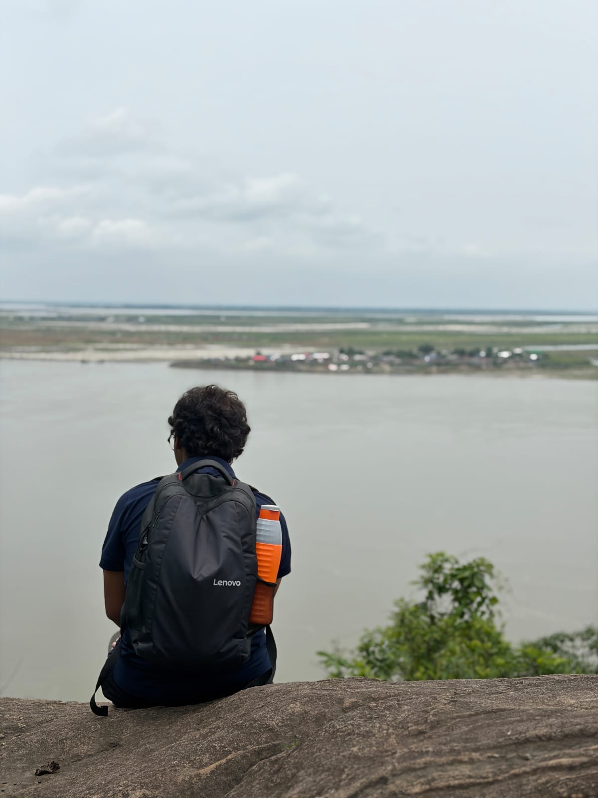 Dinesh sitting on a rock overlooking the Brahmaputra river, backpack beside him, seen from behind.